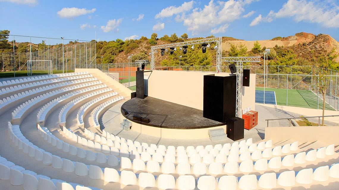 Ein Freilufttheater mit Sitzreihen und einer Bühne, umgeben von einem Zaun und einem Tennisplatz im Hintergrund.