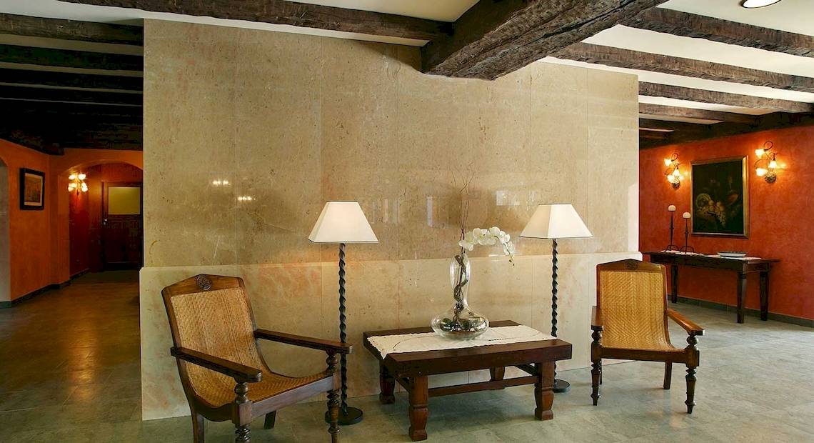 Hotellobby des Occidental Playa de Palma mit beigefarbenen Wänden, Holzbalkendecke, zwei Sesseln, einem Tisch mit Blumenvase und zwei Lampen.