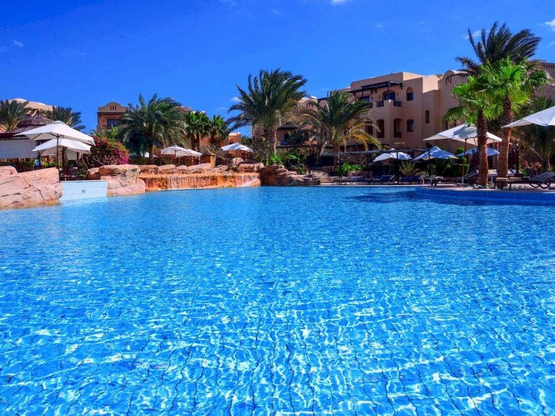 Pool im Steigenberger Coraya Beach Hotel mit Palmen und Gebäuden im Hintergrund.