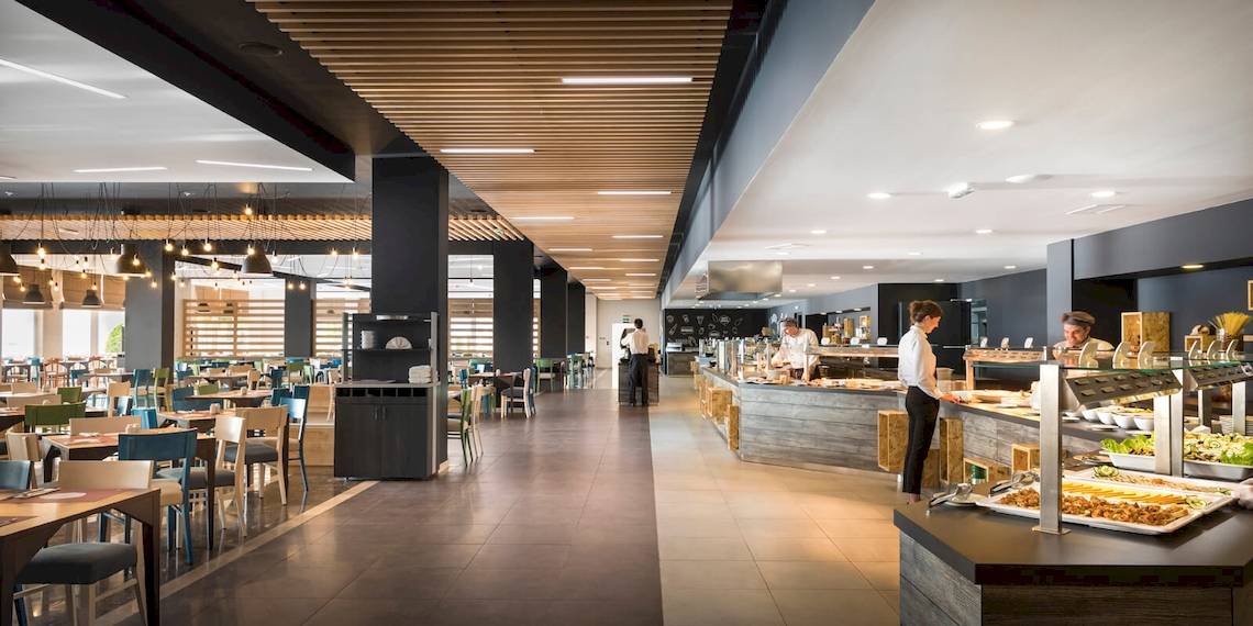 Innenansicht des Restaurants im Remisens Hotel Epidaurus mit Tischen, Stühlen und Angestellten, die Speisen zubereiten.