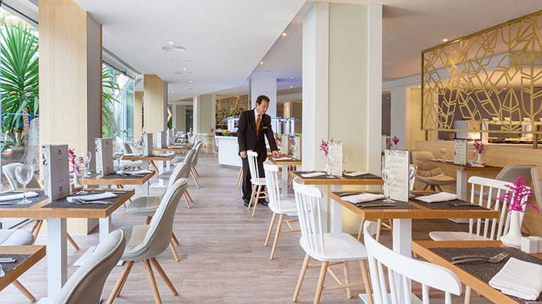 Innenansicht des Restaurants im Hotel Tigotan Lovers & Friends Playa de las Americas mit gedeckten Tischen und einem Kellner, der sich um einen Gast kümmert.