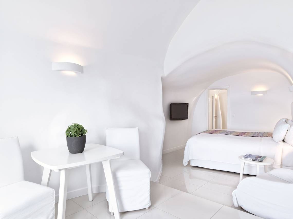 Weißes Schlafzimmer mit Bett, Tisch, Stühlen und Pflanzen im Katikies Chromata Santorini Hotel.