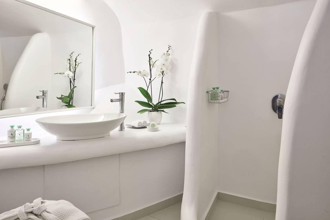 Badezimmer des Katikies Chromata Santorini Hotels mit weißem Waschbecken und Orchideen.