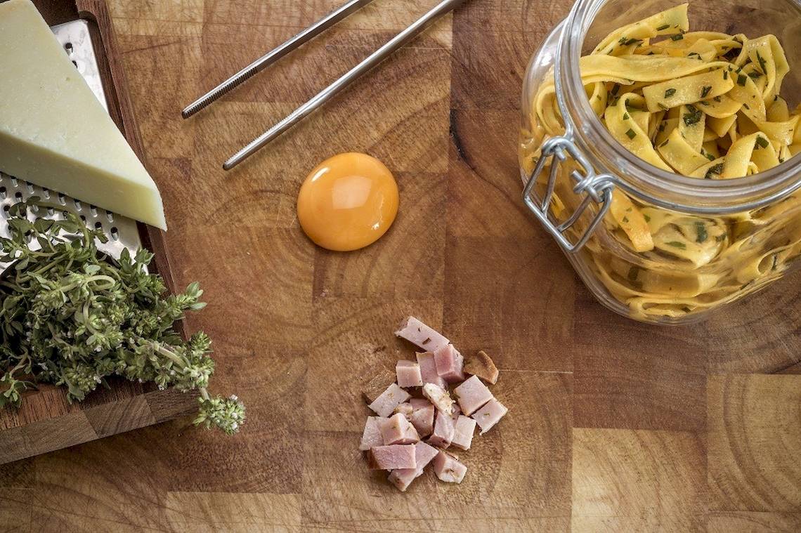 Ein Holzschneidebrett mit Zutaten für ein Rezept, einschließlich Schinken, Ei, Käse, Kräuter und Nudeln in einem Glas.