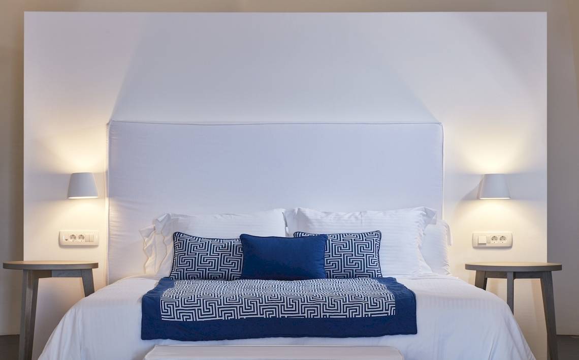 Bett im Katikies Chromata Santorini Hotelzimmer mit weißen Wänden, blauem und weißem Bettzeug und Nachttischen.