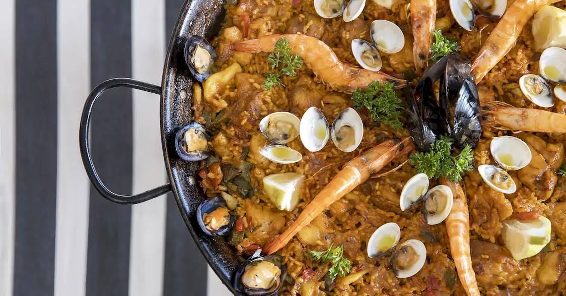 Ein großer Topf Paella mit Garnelen, Muscheln und Reis auf einem schwarzen und weißen gestreiften Hintergrund.