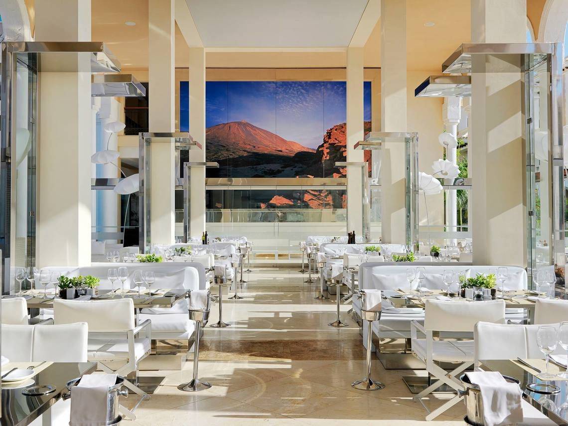 Innenbereich des Gran Melia Palacio de Isora Restaurants mit vielen weißen Tischen und Stühlen und einer großen Glaswand.