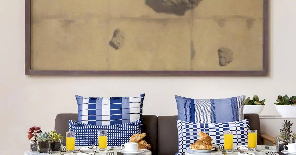 Frühstückstisch im Gran Melia Hotel mit zwei Brötchen, Orangensaft und gemusterten Kissen.