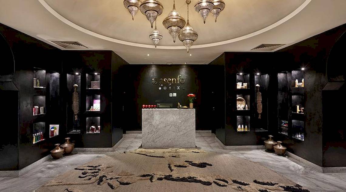 Innenbereich des Hyatt Place Taghazout Bay Hotels mit schwarzen Wänden, Marmorrezeption, hängenden Kronleuchtern und verschiedenen Regalen mit Produkten.
