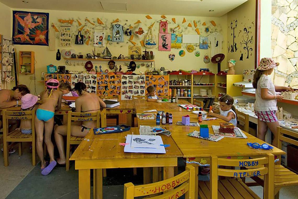 Im Blue Waters Club nehmen Kinder an einem Malkurs teil, während eine Frau im Hintergrund Bilder malt.