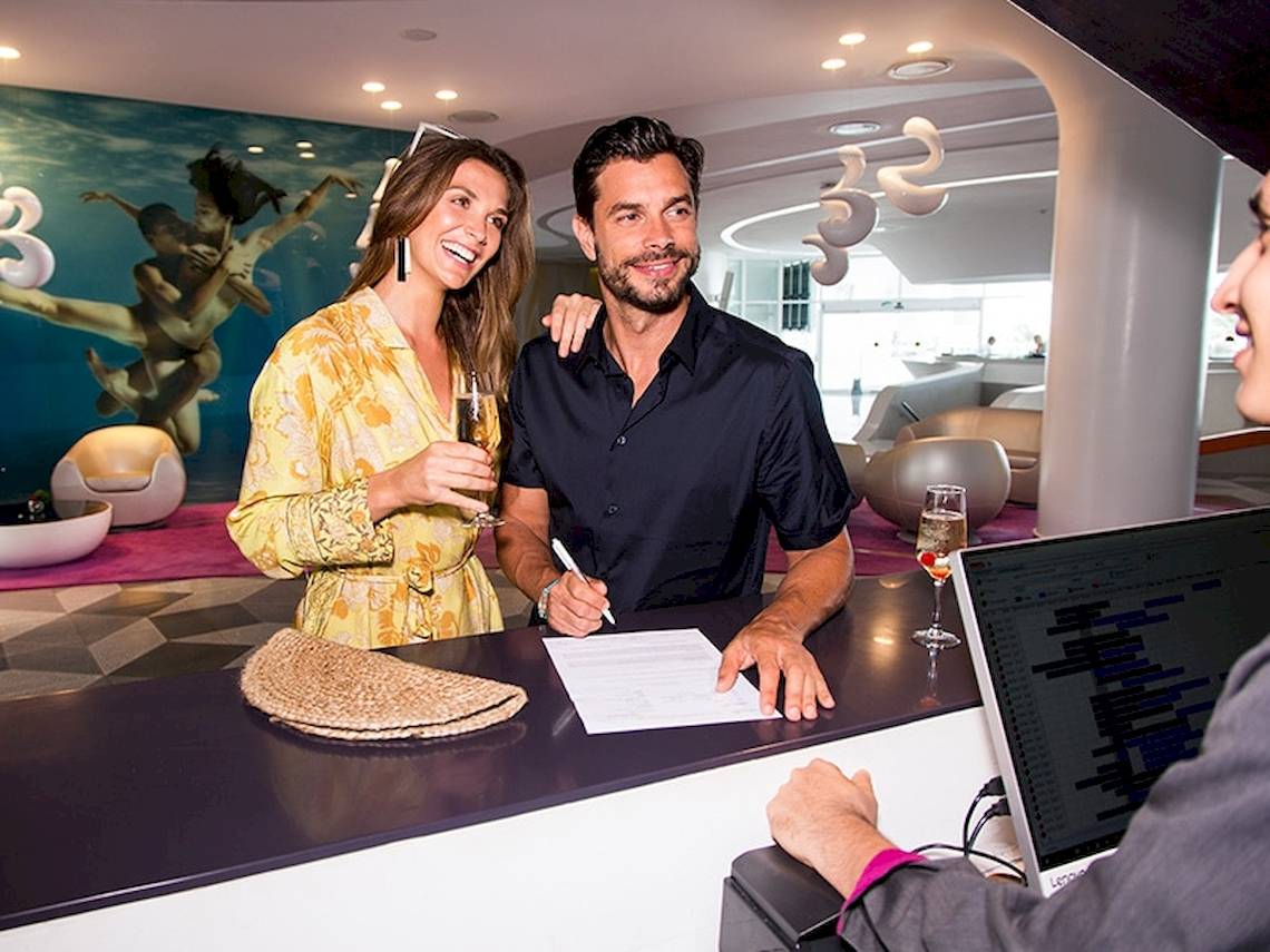 Paar im Temptation Cancun Resort Hotel, der Mann unterschreibt am Empfang, während die Frau Champagner hält.