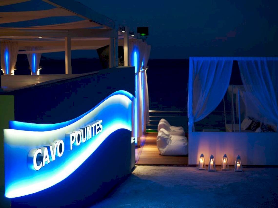 Terrasse des Proteas Blu Resorts mit blauen Lichtern und einem Schild, das 'Cavo Pountes' in weißer Schrift auf blauem Hintergrund beleuchtet.