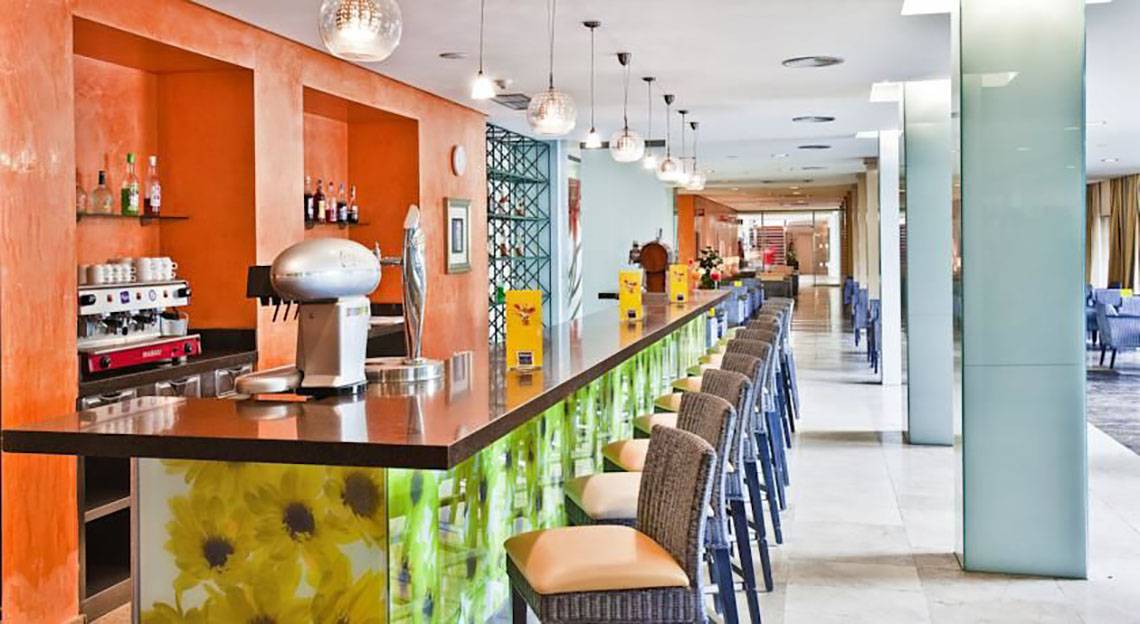 Rezeption des Barcelo Margaritas Hotels mit Bar und Stühlen, orangefarbene Wände und gelbe Blumen auf der Theke.