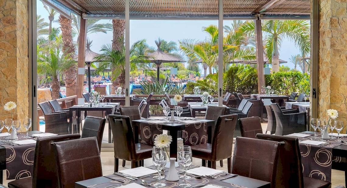 Innenansicht des Restaurants im Barcelo Fuerteventura Mar Hotel mit Tisch, Stühlen und Palmen im Hintergrund.