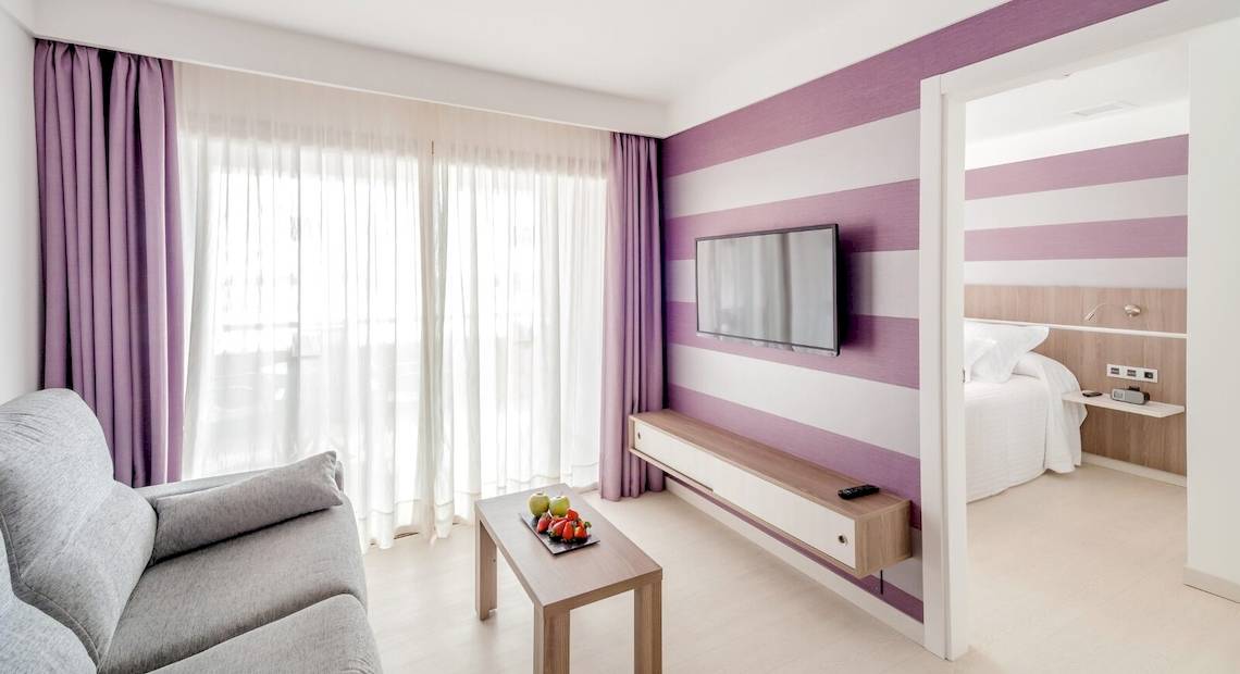 Innenansicht einer Hotelsuite im Barcelo Fuerteventura Mar mit lila und weißen Wänden, einem Sofa, einem Tisch und einem Fernseher.