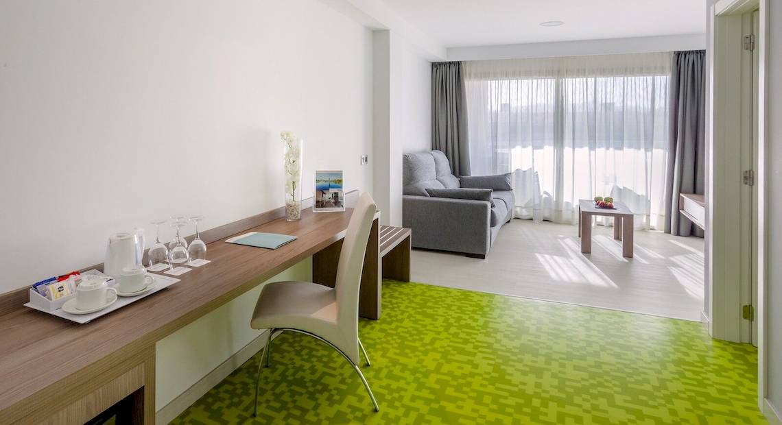 Innenansicht eines Zimmers im Barcelo Fuerteventura Mar Hotel mit Schreibtisch, Stuhl, Couch und Fenstern.