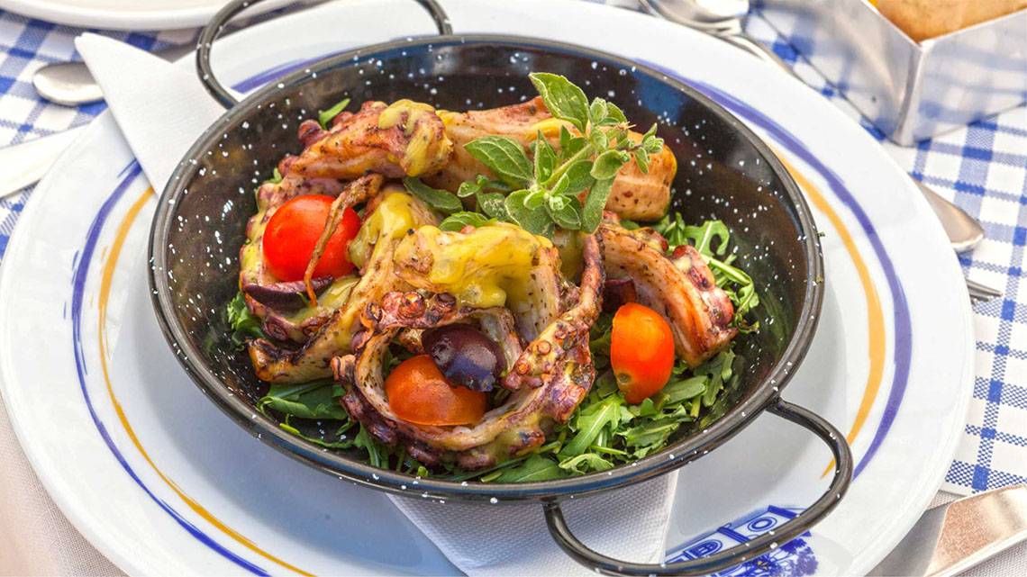 Gegrillte Oktopus-Speisen in einer schwarzen Schüssel auf einem Teller, umgeben von Salat und Tomaten.