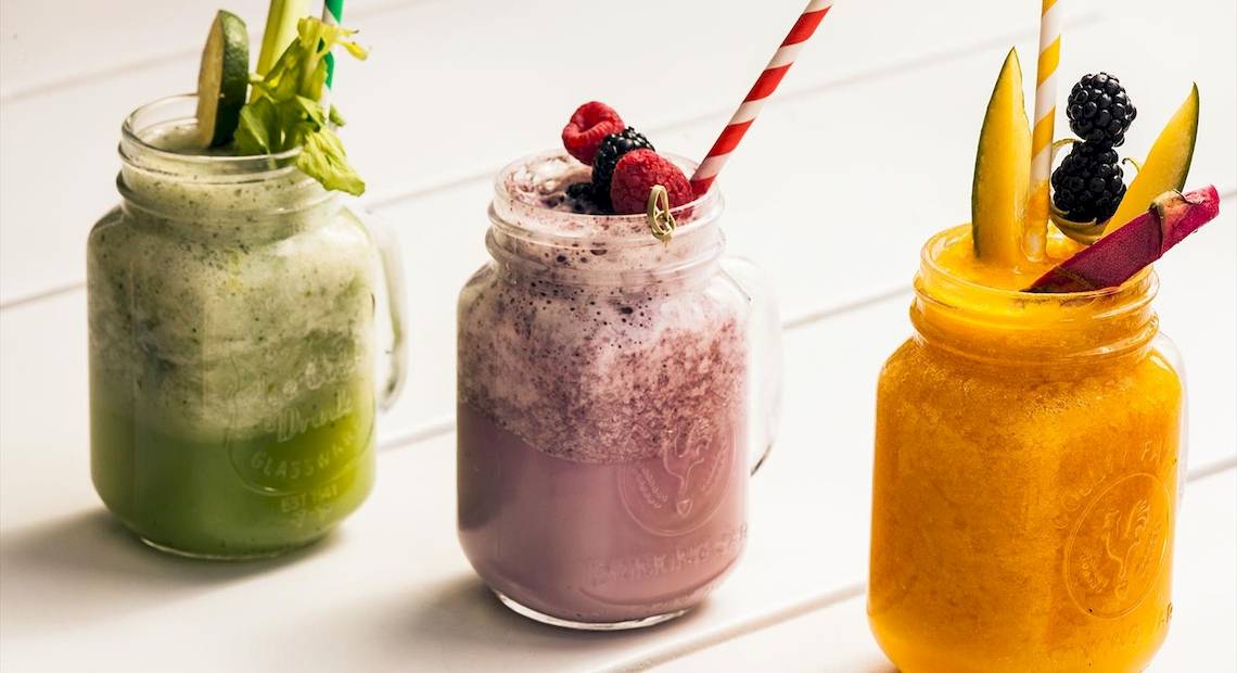 Drei Smoothie-Gläser mit verschiedenen Früchten und Strohhalmen auf einem weißen Tisch.