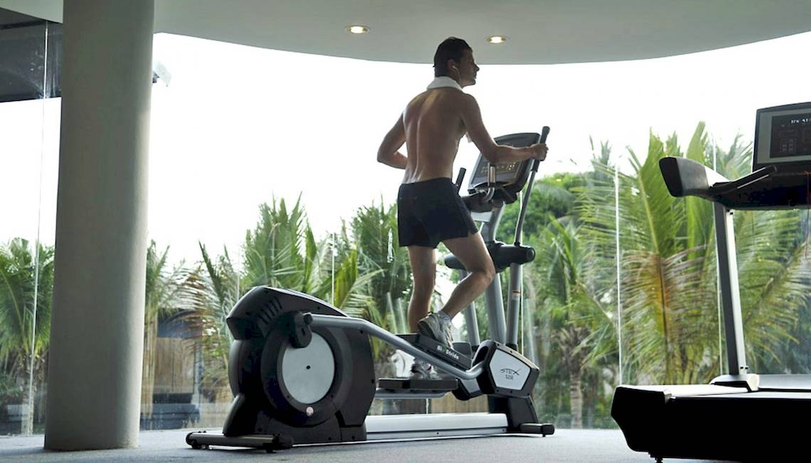 Ein Mann trainiert auf einem elliptischen Trainer im Fitnessstudio mit Blick auf Palmenbäume.