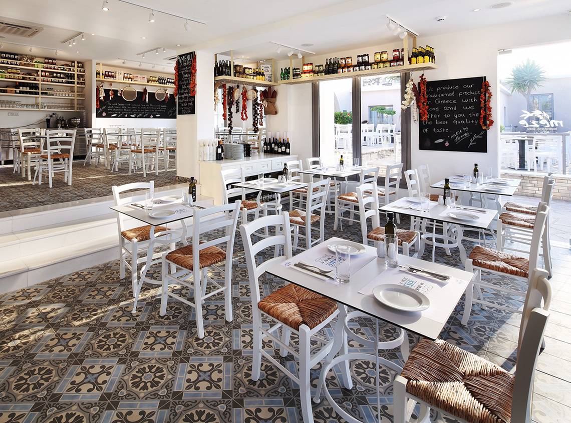 Innenansicht des Restaurants im Louis St. Elias Resort & Waterpark mit weißen Tischen und Stühlen, gefliesten Böden und einer Tafel mit Speisen.