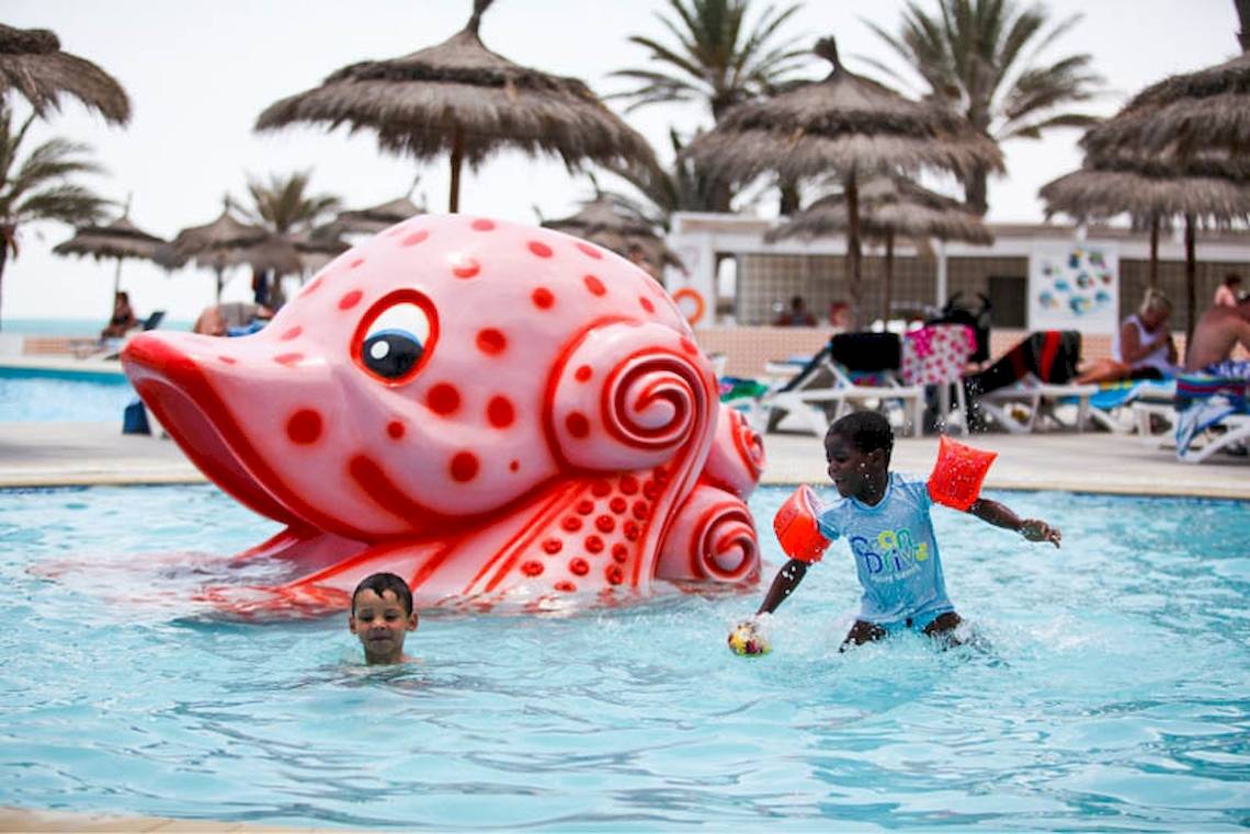 Zwei Kinder spielen im Pool mit einem großen roten Fischspielzeug im El Mouradi Djerba Menzel Hotel.