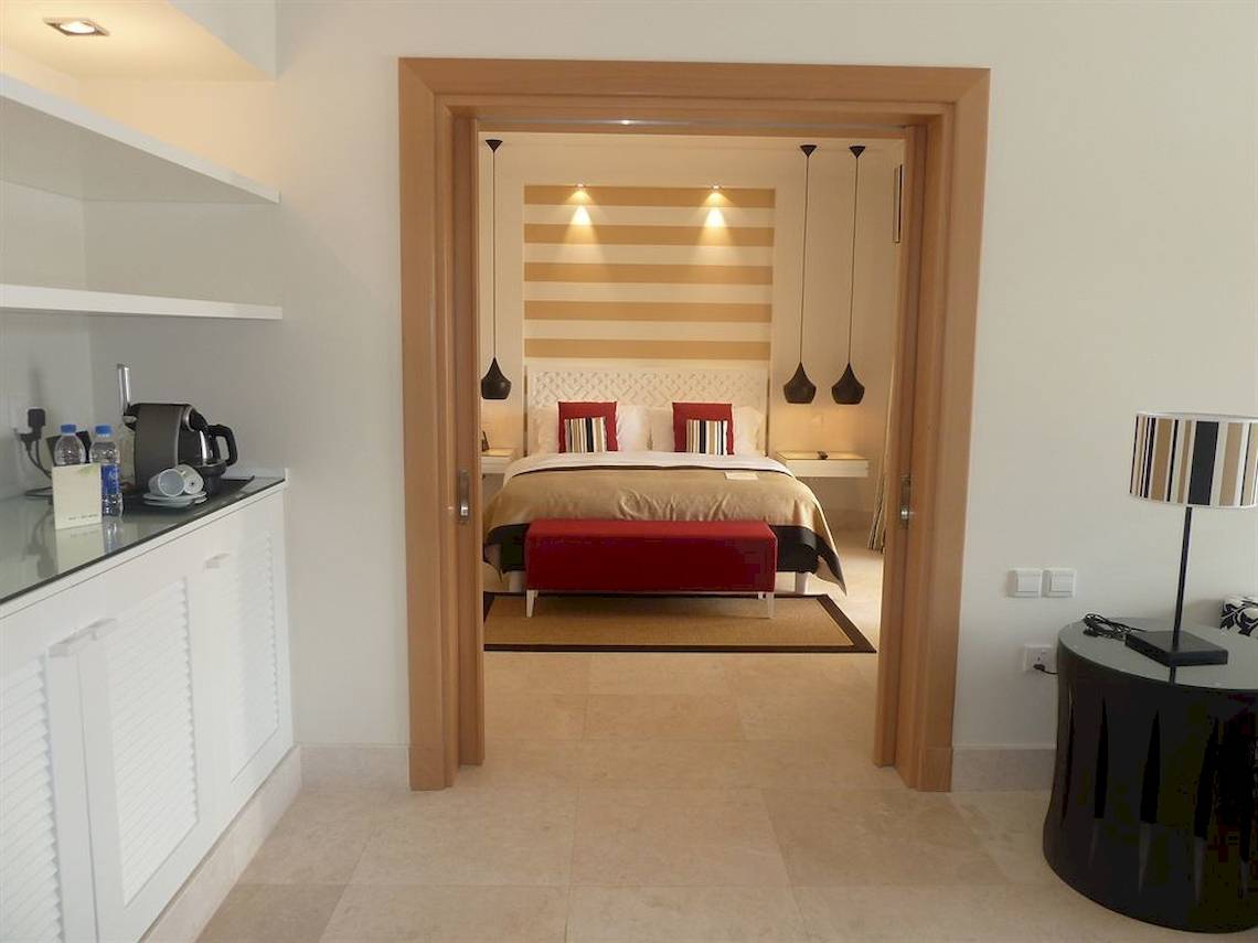 Innenansicht des Schlafzimmers im Juweira Boutique Hotel mit Doppelbett, rotem Hocker und modernen Lampen.