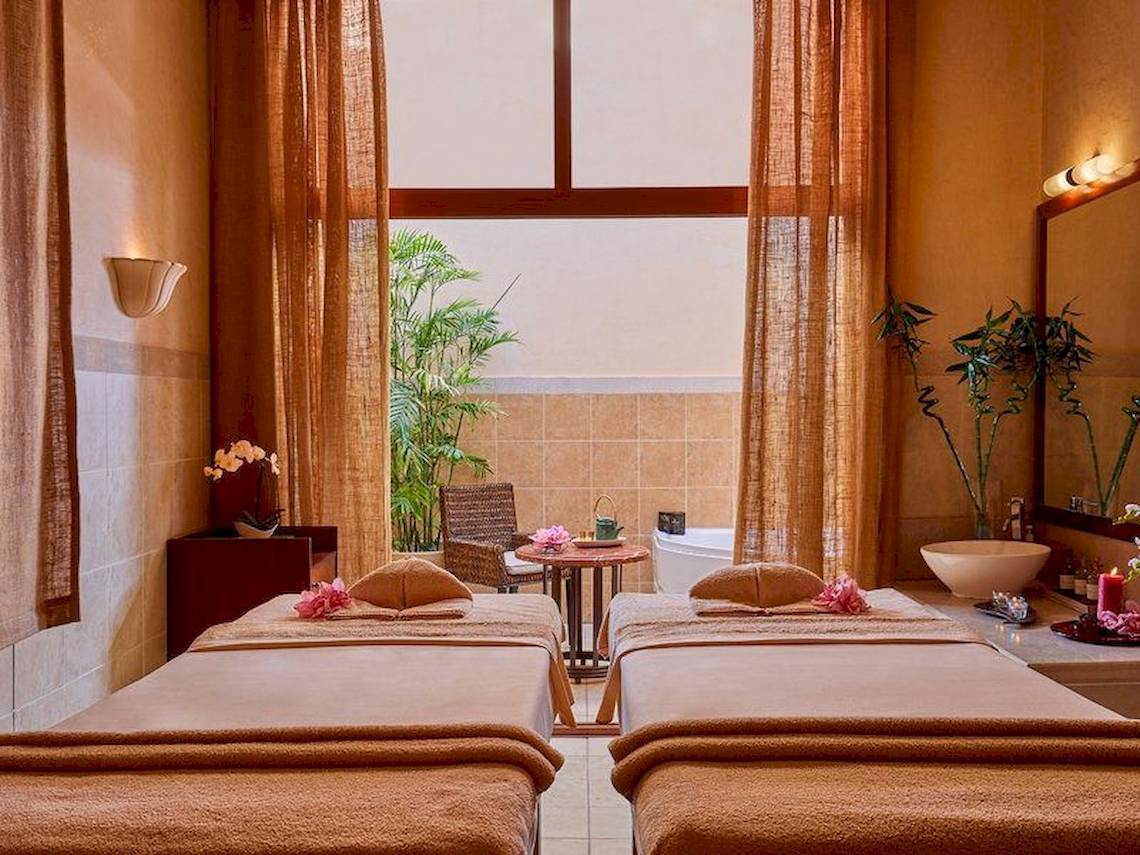 Spa-Behandlungsraum im Jaz Mirabel Beach Hotel mit zwei Massageliegen, einem Tisch und einem Fenster.