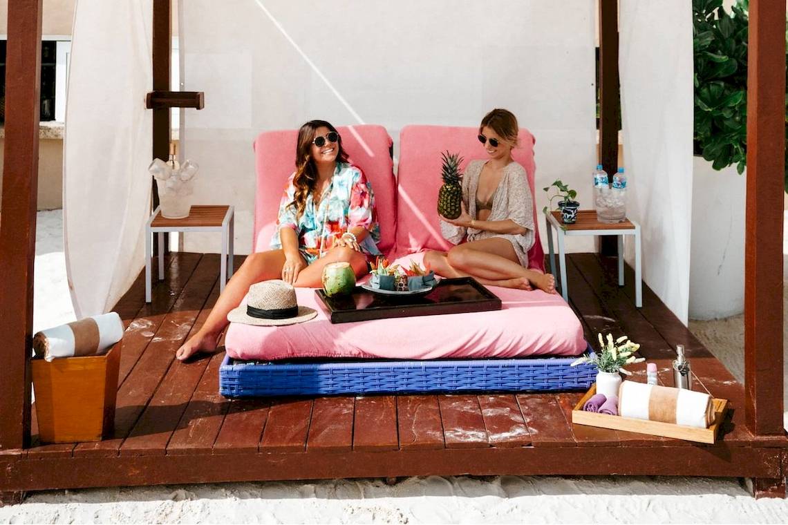 Zwei Frauen auf einem rosa Sofa sitzend auf einer Holzplattform am Strand.