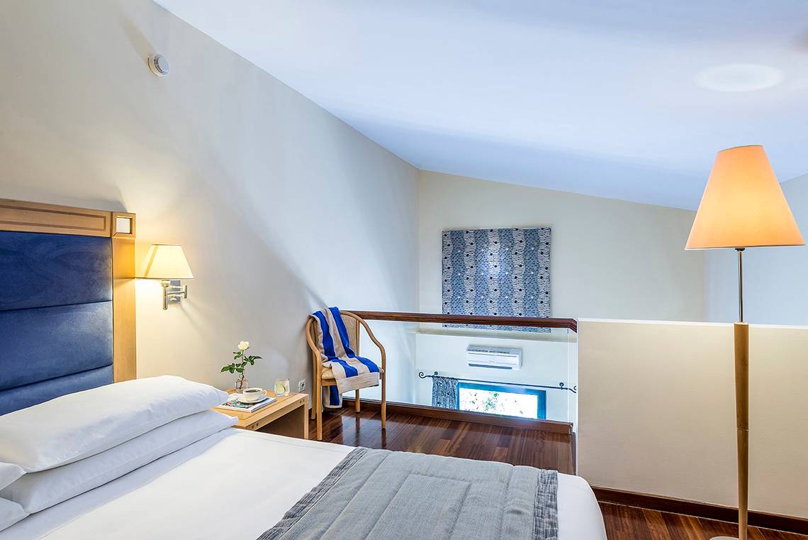 Innenansicht eines Hotelzimmers im Mitsis Lindos Memories Resort & Spa mit Bett, Nachttisch, Lampe und Balkon
