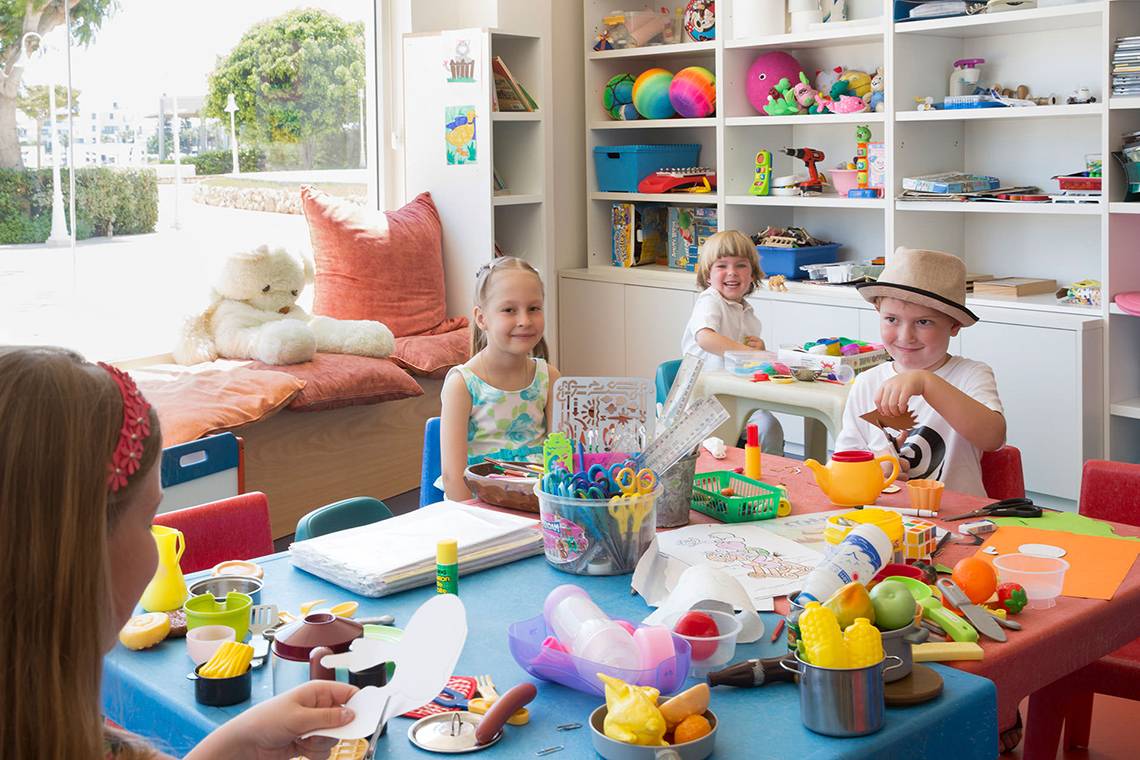 Kinder sitzen an einem Tisch im Spielzimmer des Capo Bay Hotels, umgeben von Spielzeug und Bastelmaterialien.