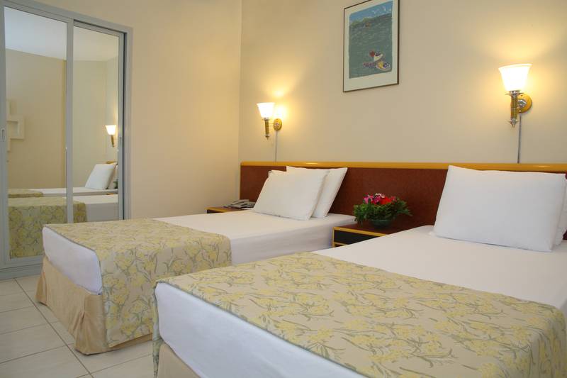 Doppelzimmer im Palm Wings Ephesus Beach Resort mit zwei Betten, weißem Bettlaken und Kissen, einem Nachttisch mit Blumen, Spiegel und Wandleuchten.