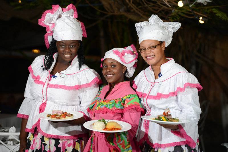 Drei Frauen in traditioneller Kleidung präsentieren verschiedene Gerichte auf Tellern.