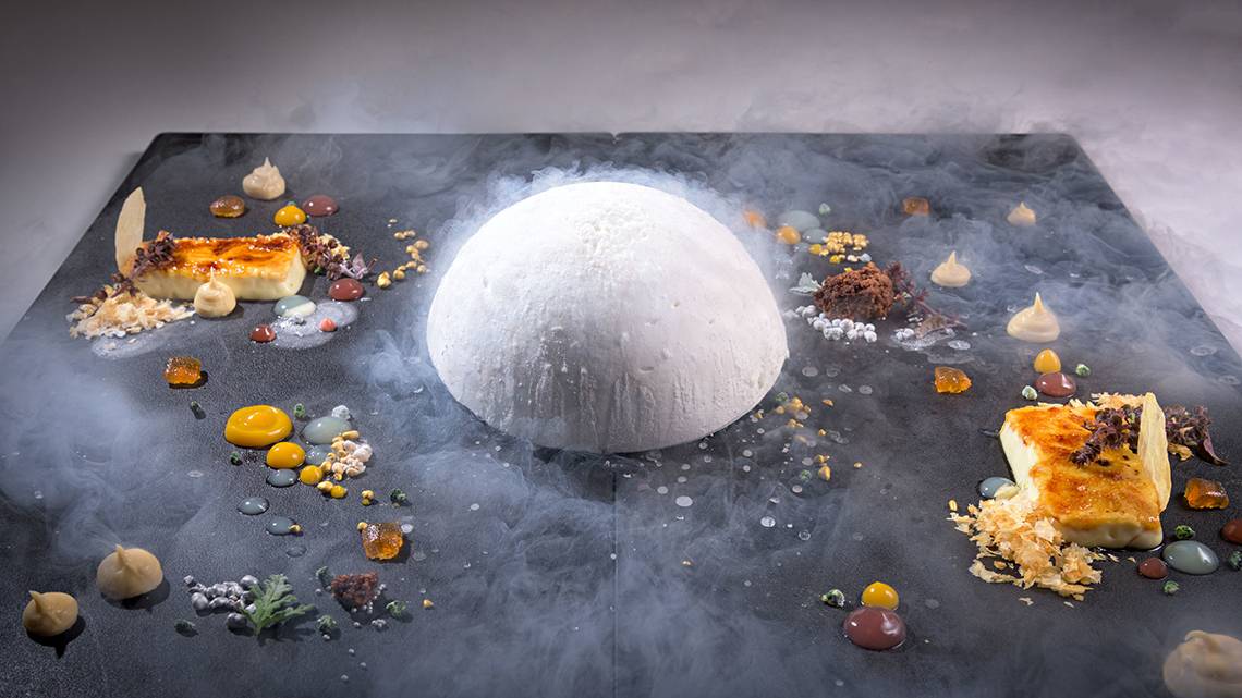Ein Dessert auf einem schwarzen Teller im Elysium Resort & Spa, umgeben von Rauch und verschiedenen Süßigkeiten.