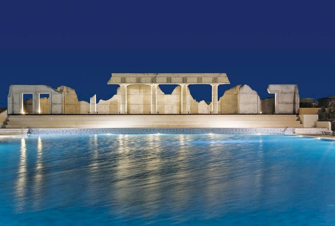 Nachtansicht des Kipriotis Village Resorts mit einem großen Pool und einer antiken Tempelarchitektur im Hintergrund.