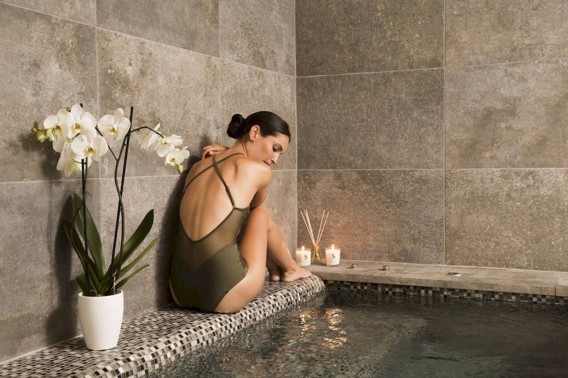 Eine Frau sitzt in einer Badewanne im Pure Salt Garonda Hotel, umgeben von weißen Orchideen, Kerzen und einer gefliesten Wand.