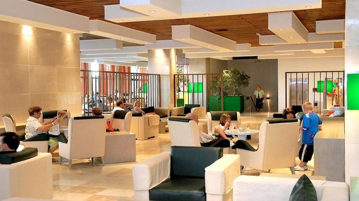 Lobby des Atlantica Nissaki Beach Hotels mit Gästen, die auf weißen Sesseln sitzen und lesen.