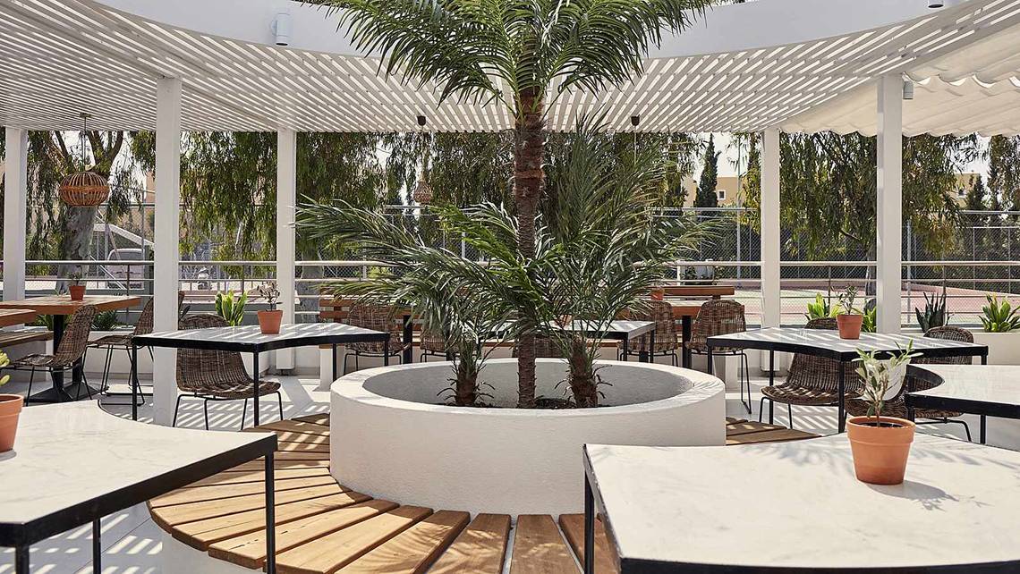 Im Freien gelegenes Restaurant des Atlantica Marmari Palace Hotels mit Tischen, Stühlen und einer großen Palme in der Mitte.