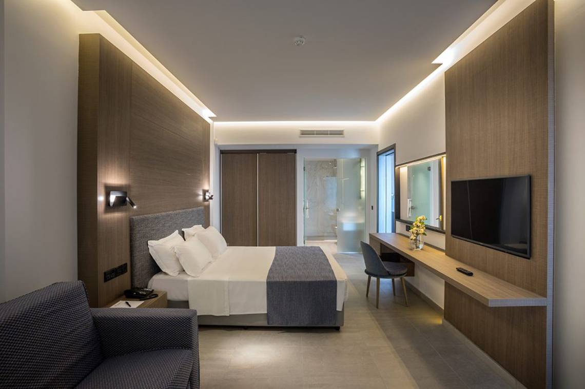 Innenansicht eines Zimmers im Cavo Maris Beach Hotel mit Bett, Schreibtisch und Fernseher.