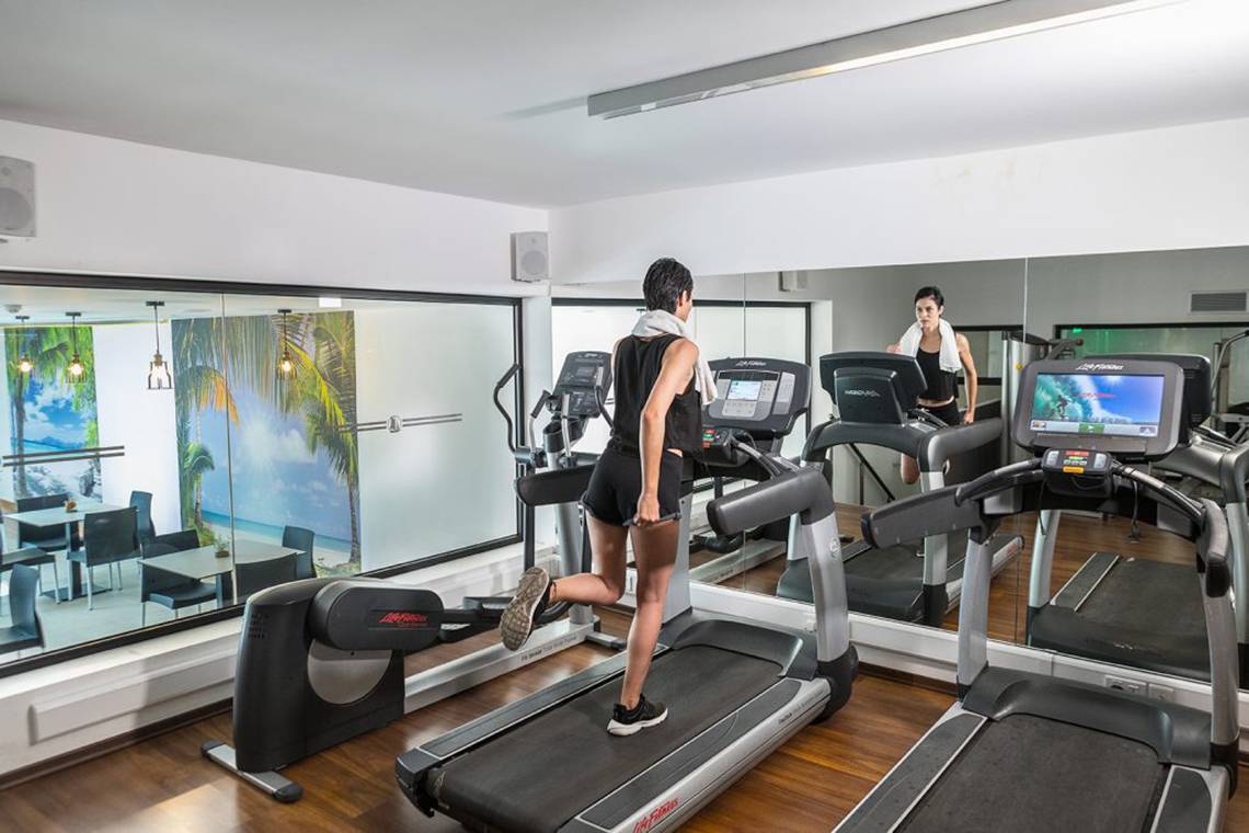 Fitnessraum im Cavo Maris Beach Hotel mit Personen auf Laufbändern und einer reflektierten Umgebung.