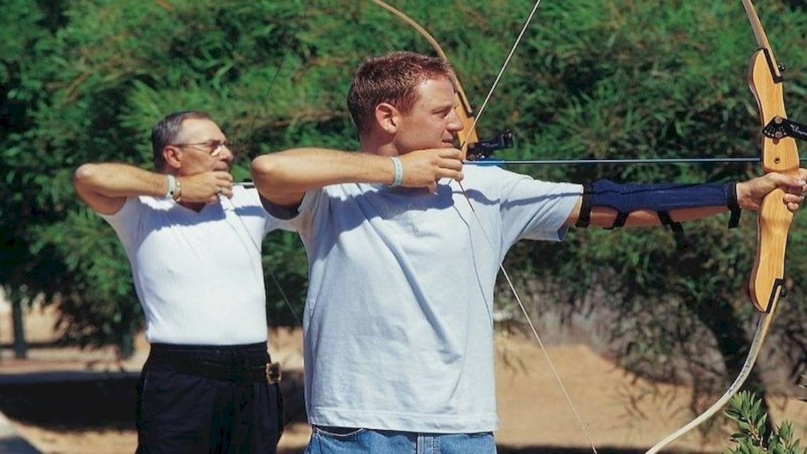 Zwei Männer schießen mit Bögen im Freien, einer mit Armschutz, vor Bäumen.