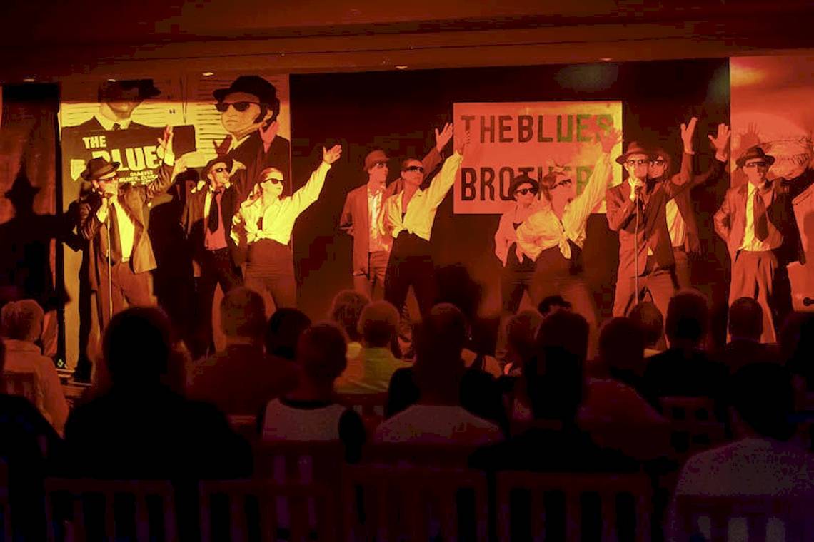 Aufführung der Blues Brothers vor Publikum auf einer Bühne mit Retro-Postern.