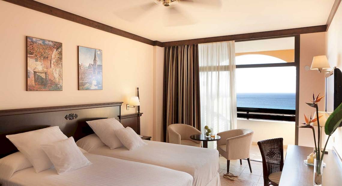 Zweibettzimmer im Occidental Jandia Royal Level mit Meerblick, zwei Betten, Sesseln, Schreibtisch und Aussicht auf das Meer.