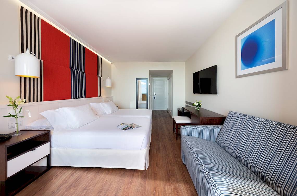Innenansicht eines Zimmers im H10 Tindaya Hotel mit zwei Betten, einer Couch und einer Fernsehwand.