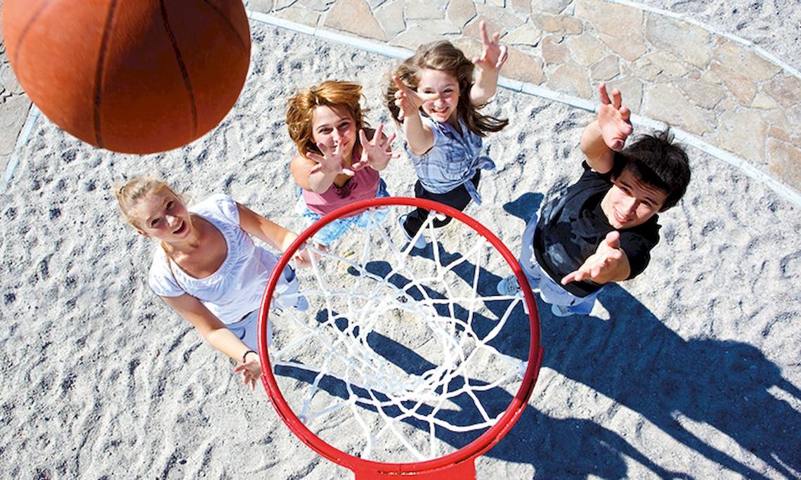 Vier Leute spielen Basketball auf Sand mit einem roten Korb und einem Basketball im Flug.