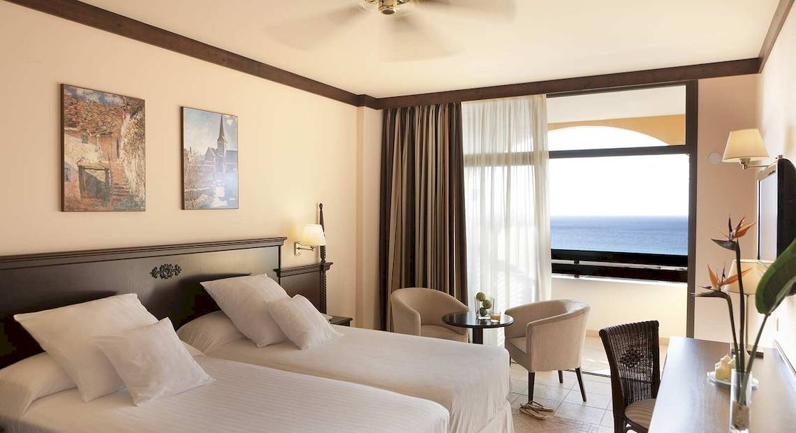 Doppelzimmer mit Meerblick im Occidental Jandia Playa Hotel, mit zwei Betten, Sesseln, einem Tisch und einem großen Fenster mit Balkon.