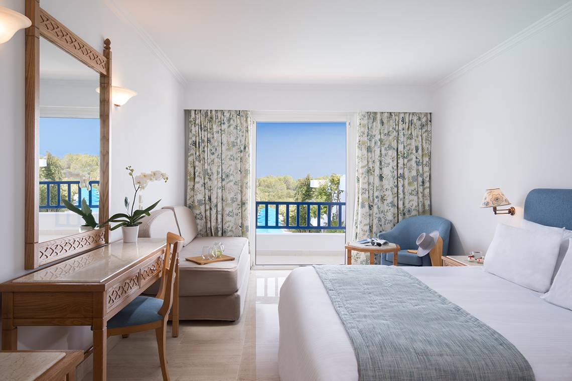 Innenansicht eines Schlafzimmers im Mitsis Ramira Beach Hotel mit Bett, Schreibtisch, Spiegel und Balkonzugang.