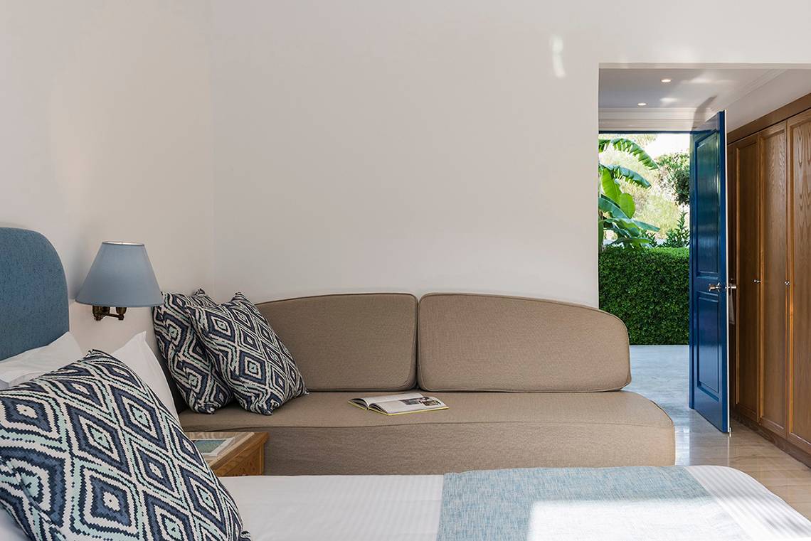 Schlafzimmer im Mitsis Ramira Beach Hotel mit Bett, Couch, Lampe und offenem Balkon.