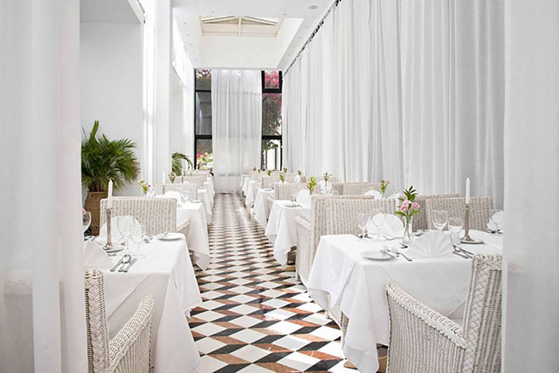 Innenansicht des Restaurants im Azia Resort & Spa mit weißen Tischen und Stühlen, gemustertem Boden und weißen Vorhängen.
