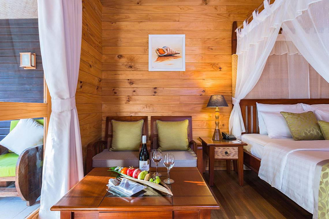 Innenansicht eines Zimmers im Bandos Maldives Hotel mit einem Bett, einem Tisch mit Obst und Weinflaschen, einem Sofa und einem Gemälde an der Wand.