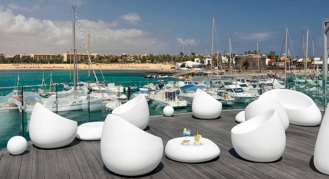 Terrasse des Barcelo Fuerteventura Castillo mit weißen Stühlen und Tischen, Blick auf den Hafen mit vielen Booten.